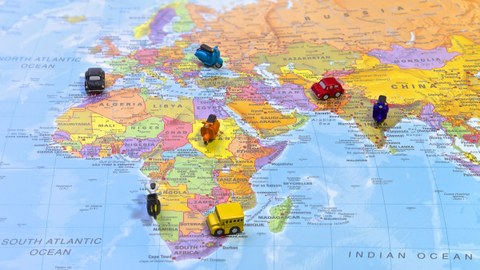 Bild einer Weltkarte. Es sind nur die Kontinente Afrika, Asien und Europa zu sehen. Sieben Miniatur-Fahrzeuge (Auto, Bus, Roller) stehen verteilt auf der Karte.