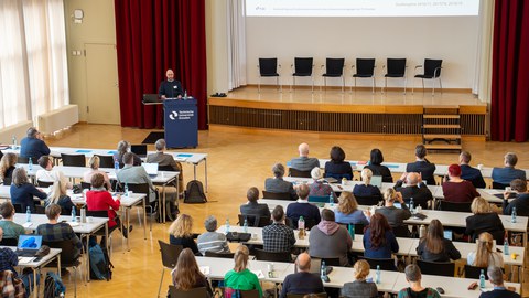 Foto: In einem Saal steht eine Person vor Publikum am Rednerpult.
