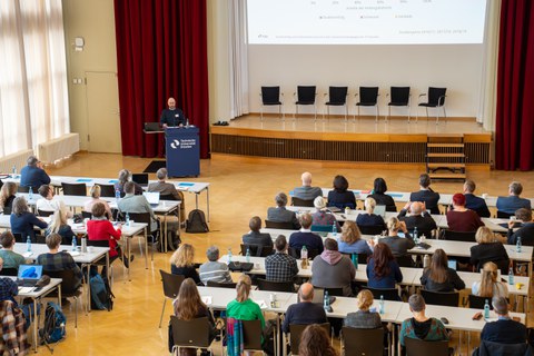 Foto: In einem Saal steht eine Person vor Publikum am Rednerpult.