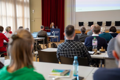 Foto: In einem Saal steht eine Person vor Publikum am Rednerpult.