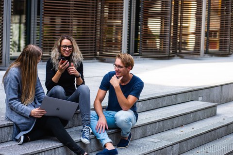 Bild einer Gruppe von 3 Personen, die zusammen vor einem Gebäude auf einer Treppe sitzen und sich miteinander unterhalten 