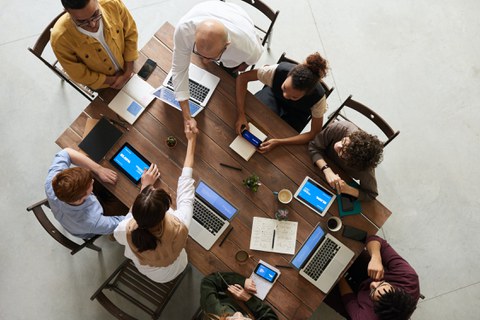 Foto einer Gruppe von 8 Personen in Vogelperspektive. Die Personen sitzen um einen Holztisch und arbeiten an mobilen Endgeräten, Laptops oder mit Notizbüchern. Eine Frau und ein Mann reichen sich über den Tisch hinweg die Hand.