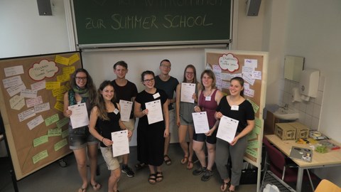 Bild von 8 Studierenden, die in einem Seminarraum vor einer Tafel stehen, auf der "SummerSchool" steht. Sie halten jeweils eine Urkunde in ihren Händen und lächeln in die Kamera.