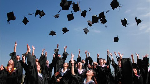 Das Foto zeigt eine Gruppe von Absolventen in langen schwarzen Mänteln. Sie werfen zeitgleich ihre Doktorhüte in die Luft und lachen dabei.