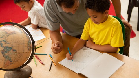 Das Foto zeigt zwei Kinder, die an einem Tisch sitzen und mit Farbstiften etwas in ein leeres Heft malen. Hinter dem einen Kind kniet ein Mann und schaut dem Kind über die Schulter. Auf dem Tisch befindet sich ebenfalls ein Globus.