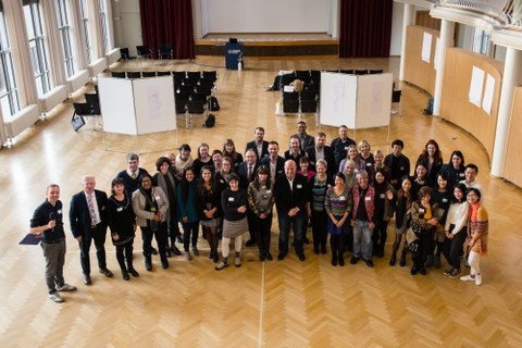 Bild von einer Gruppe aus über 20 Personen, die in einem ansonsten leeren Saal mit Stuhlreihen und einer Bühne stehen und in die Kamera lächeln.