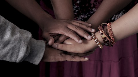 Das Foto zeigt die Hände mehrerer Personen. Sie stehen im Kreis und haben die Hände übereinander gelegt. Eine Person trägt Armschmuck.