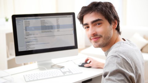 Bild eines Mannes, der sich über die linke Schulter in Richtung Kamera dreht. Er sitzt vor einem Computer und bedient mit der rechten Hand eine Maus. 