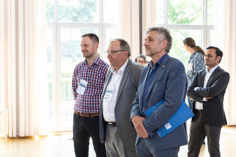 Das Foto zeigt drei Männer, die nebeneinander stehen und bei einer Vorstellungsrunde zuhören.