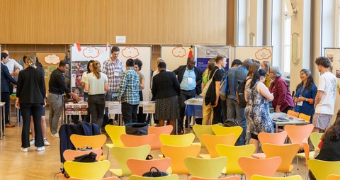 Das Foto zeigt reges Treiben von Leuten aus verschiedenen Ländern vor Moderationswänden.  Im Vordergrund sind mehrere Stuhlreihen zu sehen.