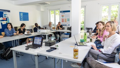 Das Foto zeigt einen Raum mit ca. 10 Personen während eines Workshops. Dabei sind die Zuhörer:innen von vorne fotografiert.