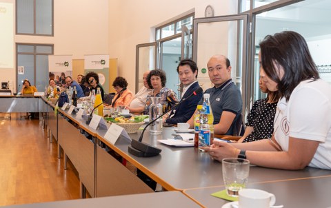 Das Foto zeigt mehrere Personen, die in einer Reihe an einem Konferenztisch sitzen mit Mikrofon an jedem Platz.