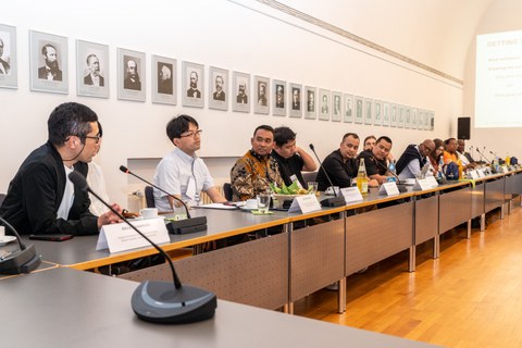Das Foto zeigt mehrere Personen, die in einer Reihe an einem Konferenztisch sitzen mit Mikrofon an jedem Platz.