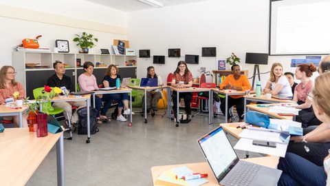 Das Foto zeigt eine Gruppe von ca. 10 Personen, die auf Bestuhlung in U-Form sitzen und an einem Workshop teilnehmen