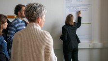 Foto: Mehrere Personen stehen vor einem Plakat, das an einer Wand befestigt ist. Eine Person schreibt etwas auf das Plakat.