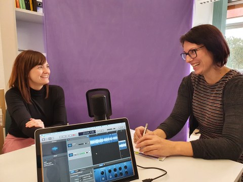 Auf dem Foto sind zwei Frauen zu sehen, die an einem Tisch sitzen, sich anschauen und lachen. Auf dem Tisch im Vordergrund sind ein Laptop und ein Podcastmikrofon zentral platziert. Im Hintergrund steht eine lila Wand.