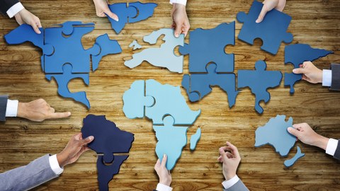 Foto. Auf einem Holzuntergrund liegen Puzzle-Teile, welche die Kontinente der Welt formen. 8 Hände halten jeweils ein Puzzleteil. 2 Hände zeigen auf das Puzzle.