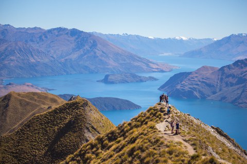 Das Foto zeigt eine bergige Landschaft, in der mehrere Personen wandern und sich fotografieren. Im Hintergrund des Bildes ist ein großer See zu sehen.