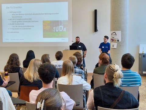Zwei Referenten stehen vor einer sitzenden Zuhörerschaft an einem Rednerpult und präsentieren eine Folie mit dem Titel 'Die TU Dresden'. Die Folie listet Eckdaten auf: 1828 gegründet, seit 2012 Exzellenzuniversität, 29.000 Studierende, davon 14,3 % Lehram