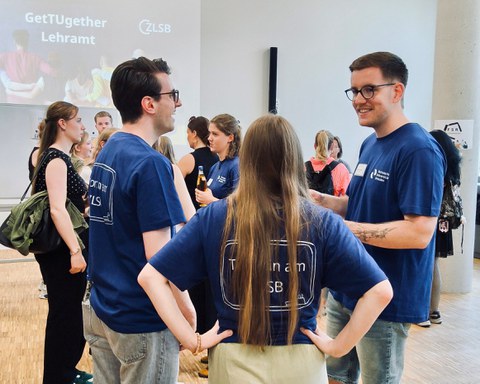 Drei Personen in blauen T-Shirts mit der Aufschrift 'Tutor:innen am ZLSB' unterhalten sich im Kreis stehend, im Hintergrund läuft eine Präsentation mit dem Titel 'GetTUgether Lehramt' und weitere Gäste stehen im Gespräch beisammen.