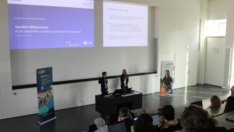 Foto: zwei Personen stehen in einem Hörsaal hinter dem Redner:innenpult.