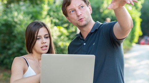 Foto von zwei Personen im Freien, die ein Laptop halten. Der Mann zeigt auf etwas vor ihnen.