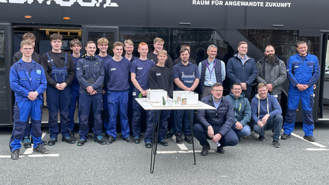 Foto: Gruppenbild von 20 Männern in blauer Arbeitskleidung vor einem schwarzen Omnibus mit der Aufschrift "Fabmobil. Raum für angewandte Zukunft".