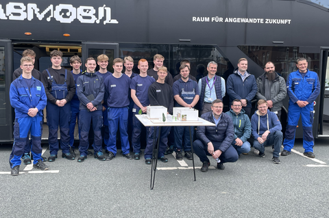 Foto: Gruppenbild von 20 Männern in blauer Arbeitskleidung vor einem schwarzen Omnibus mit der Aufschrift "Fabmobil. Raum für angewandte Zukunft".