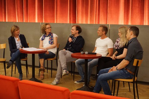 Bild einer Gesprächsrunde von 6 Personen, die im Programmkino Ost um zwei Tische herum sitzen. Ein Mann hat ein Mikrofon in der Hand und redet. Zwei weitere Mikrofone liegen auf dem rechten Tisch.