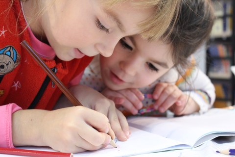 Bild von zwei kleinen Kindern, die sich gemeinsam über ein Heft beugen, in das eine der beiden schreibt.