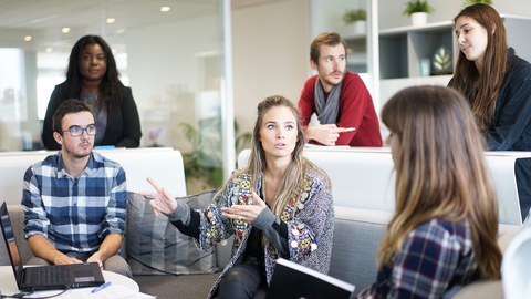 Bild von 6 Personen. 3 Menschen sitzen um einen Tisch auf einer Couch und reden miteinander. Die 3 anderen Personen stehen hinter der Couch und hören zu.