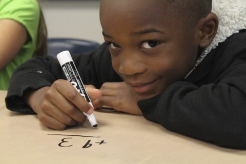 Bild eines Jungen, der mit einem schwarzen Stift auf Papier schreibt und in die Kamera lächelt.