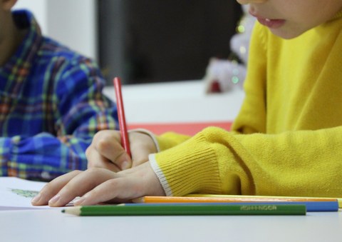 Bild von 2 Kindern, die an einem Tisch sitzen. Das vordere Kind malt mit einem roten Buntstift in ein Heft. Neben dem Heft liegen weitere Buntstifte in verschiedenen Farben.