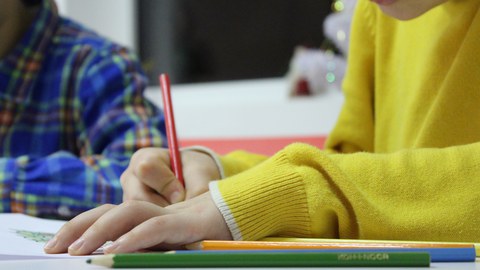 Bild von 2 Kindern, die an einem Tisch sitzen. Das vordere Kind malt mit einem roten Buntstift in ein Heft. Neben dem Heft liegen weitere Buntstifte in verschiedenen Farben.