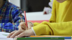 Bild von 2 Kindern, die an einem Tisch sitzen. Das vordere Kind malt mit einem roten Buntstift in ein Heft. Neben dem Heft liegen weitere Buntstifte in verschiedenen Farben.