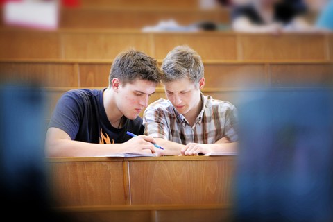 Bild eines Hörsaals. Im Mittelpunkt sind 2 männliche Studenten, die nebeneinander sitzen und gemeinsam in die Unterlagen schauen. Im Hintergrund sind weitere Studierende zu erkennen.