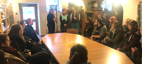 Bild einer Gruppe von Menschen, die in der Schkola Grund- und Oberschule Ostritz um eine Tafel sitzen und 3 Frauen zuhören, die im Hintergrund stehen.