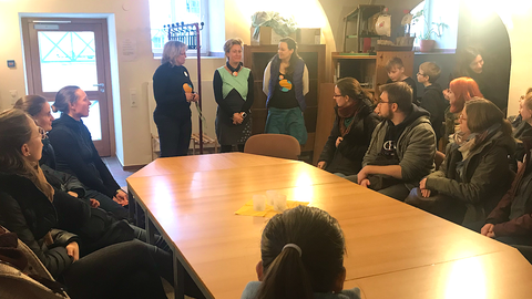 Bild einer Gruppe von Menschen, die in der Schkola Grund- und Oberschule Ostritz um eine Tafel sitzen und 3 Frauen zuhören, die im Hintergrund stehen.