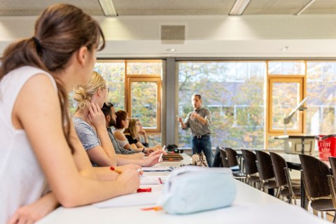 Foto: Eine Gruppe von Personen sitzt in einem Seminarraum und hört einer Lehrperson zu.