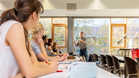 Foto: Eine Gruppe von Personen sitzt in einem Seminarraum und hört einer Lehrperson zu.