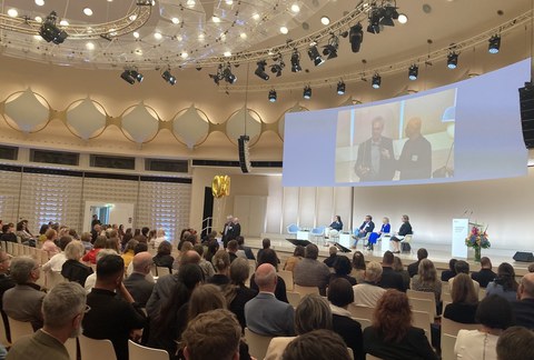 Foto: Blick in einen Veranstaltungssaal mit zahlreichen Besucher:innen