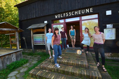 Foto: 6 Personen in Sommerkleidung stehen vor dem Eingang zu einem hölzernen Gebäude. Über der Eingangstür ist die Aufschrift "Wolfsscheune" angebracht.