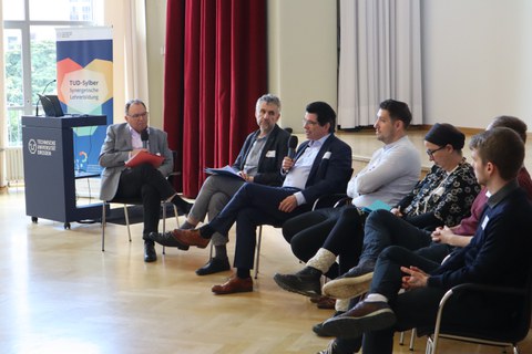 Foto: 7 Personen sitzen in einer Stuhlreihe vor einer Bühne.