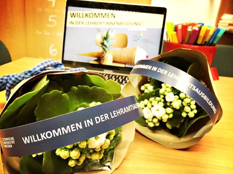 Foto: auf einem Tisch liegen kleine Blumensträuße, auf deren Verpackung "Willkommen in der Lehramtsausbildung" steht. Im Hintergrund ist ein Laptop zu sehen, auf dessen Bildschirm der Text "Willkommen in der Lehramtsausbildung" zu sehen ist.