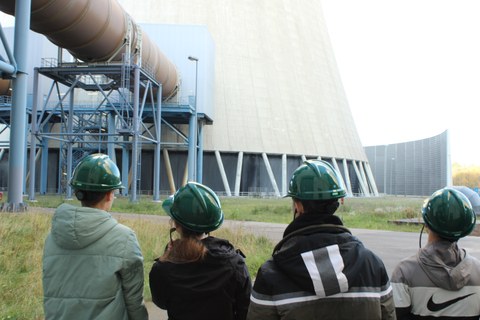 Foto: Rückenansicht von vier Personen mit grünen Schutzhelmen, die auf eine technische Anlage blicken.