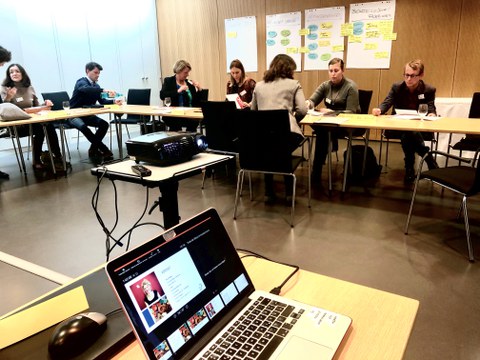 Bild eines Seminarraumes, in dem 7 Personen an verschiedenen Tischen sitzen. Im Vordergrund steht ein Laptop auf einem Tisch. Er ist mit einem Beamer verbunden.