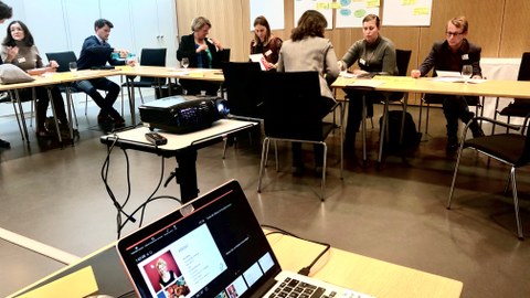 Bild eines Seminarraumes, in dem 7 Personen an verschiedenen Tischen sitzen. Im Vordergrund steht ein Laptop auf einem Tisch. Er ist mit einem Beamer verbunden.