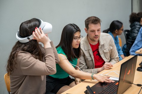 Foto: Drei Personen sitzen nebeneinander an einem Tisch. Zwei blicken gemeinsam auf den Bildschirm eines Laptops, eine trägt eine VR-Brille.