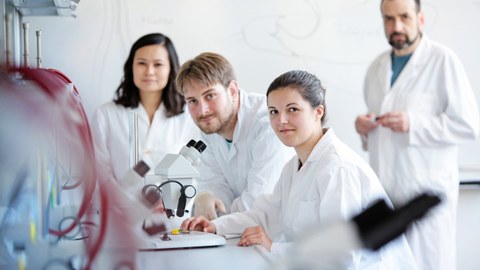 Foto: Mehrere Personen in weißer Laborkleidung stehen um ein Mikroskop