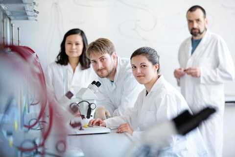 Foto: Mehrere Personen in weißer Laborkleidung stehen um ein Mikroskop